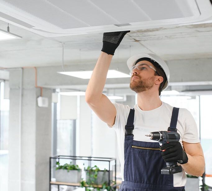man holding a hand drill while inspecting a ceiling mounted air conditioning system